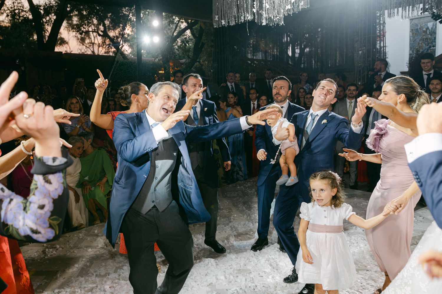 Wedding party dances energetically on reception dance floor with hanging decor above.