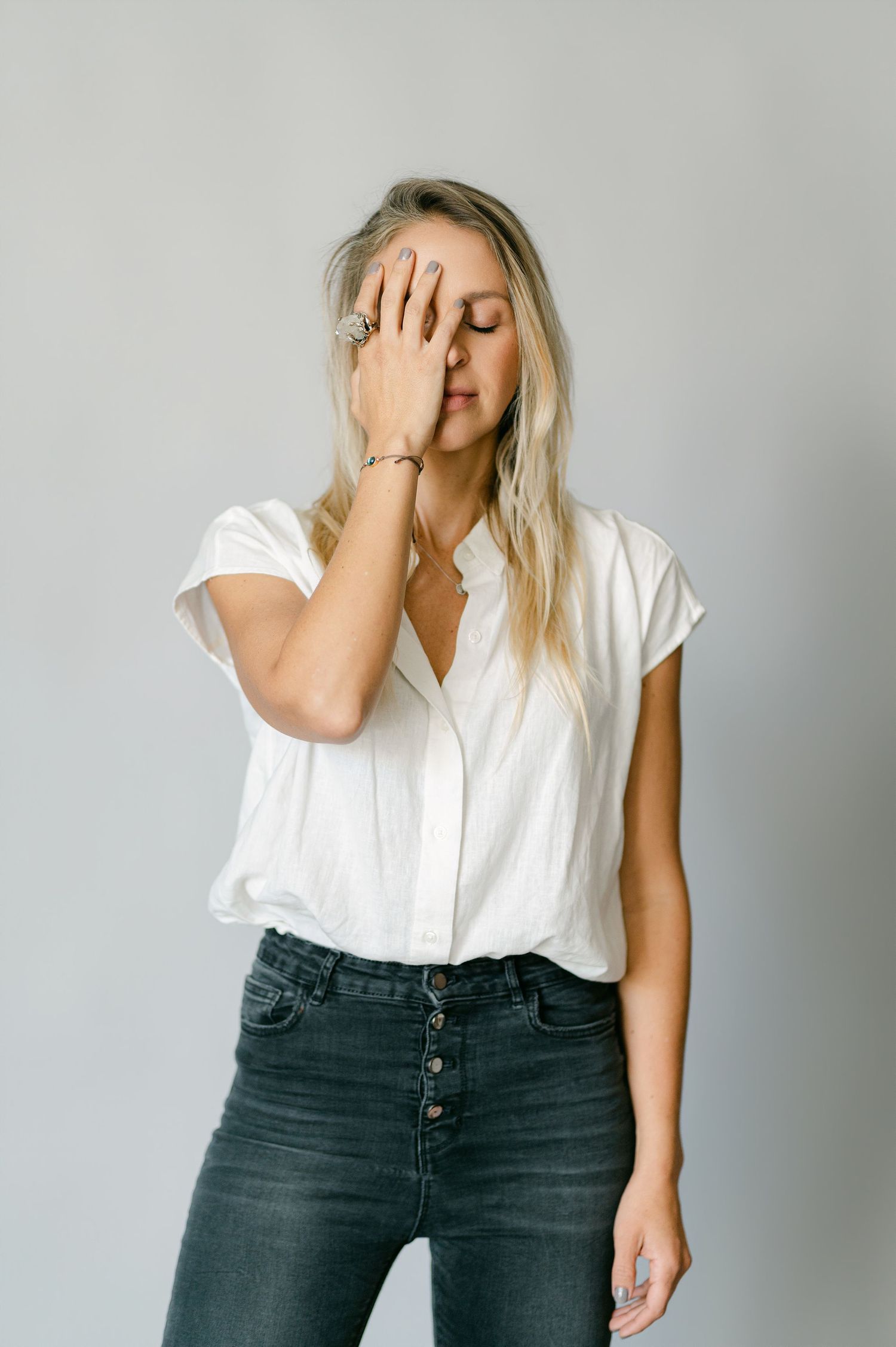Blonde woman in white blouse and dark jeans covering face with hand, studio portrait.