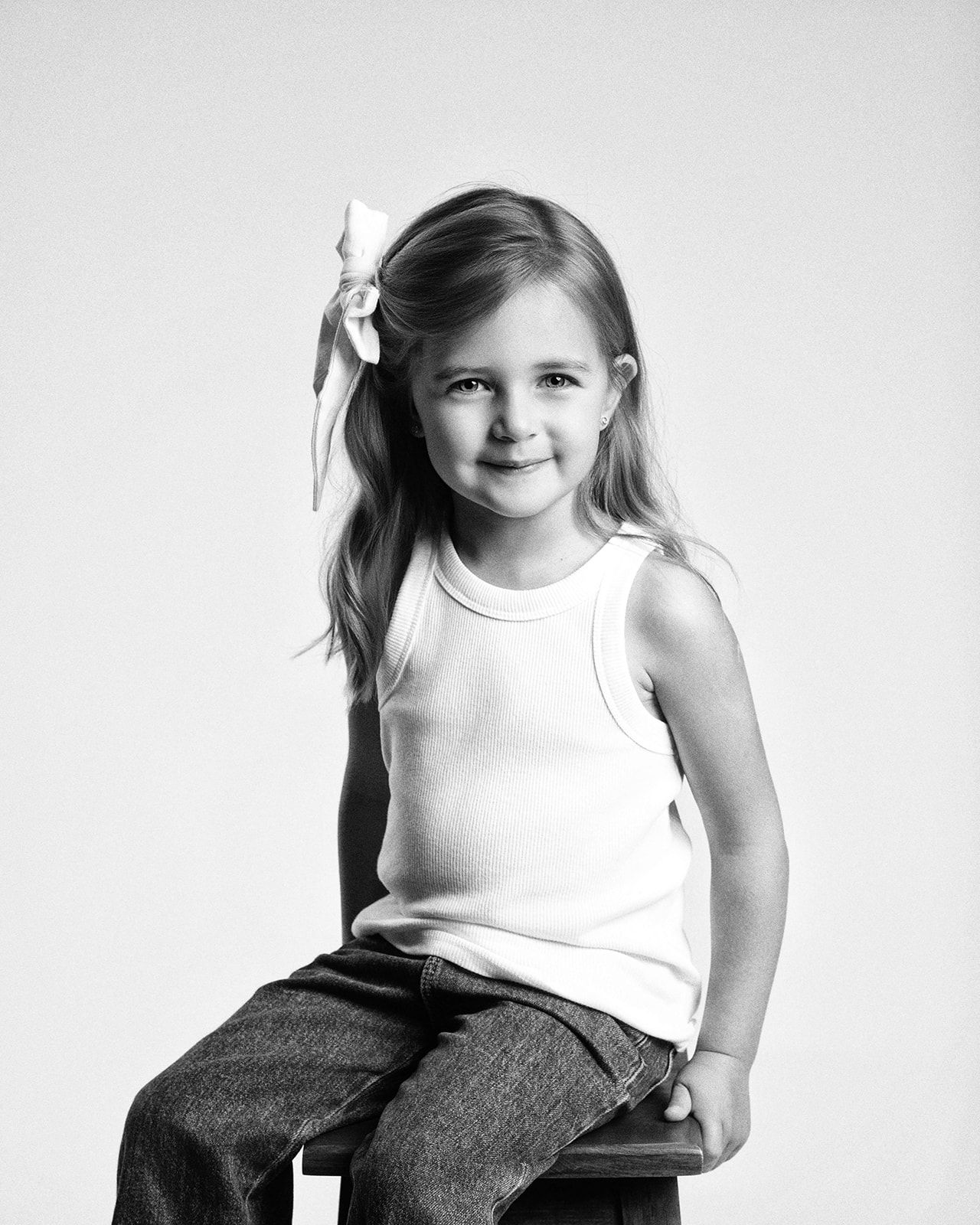 A young girl with a bow in her hair sits on a stool in a white tank top and jeans in a B&W portrait.