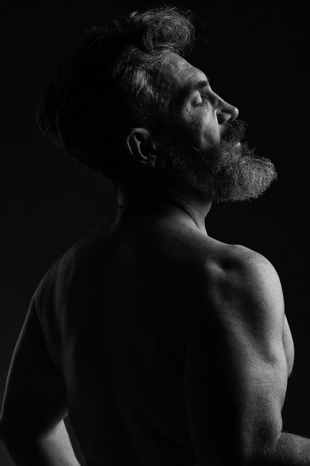 Black and white portrait of a shirtless bearded man with eyes closed, facing sideways against dark background.
