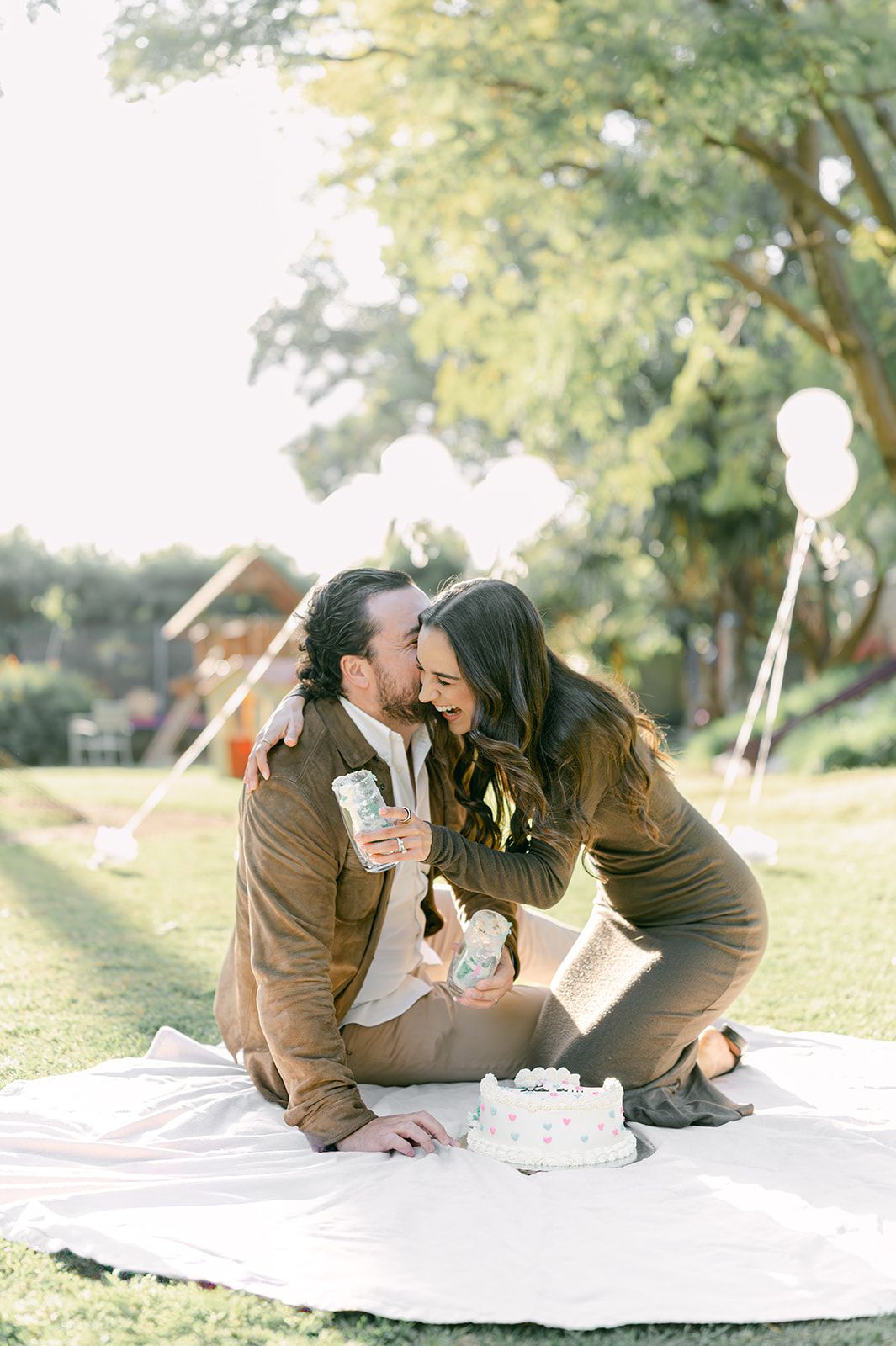 Joyful expecting couple kissing on picnic blanket with gender reveal cake and balloons in sunny park.