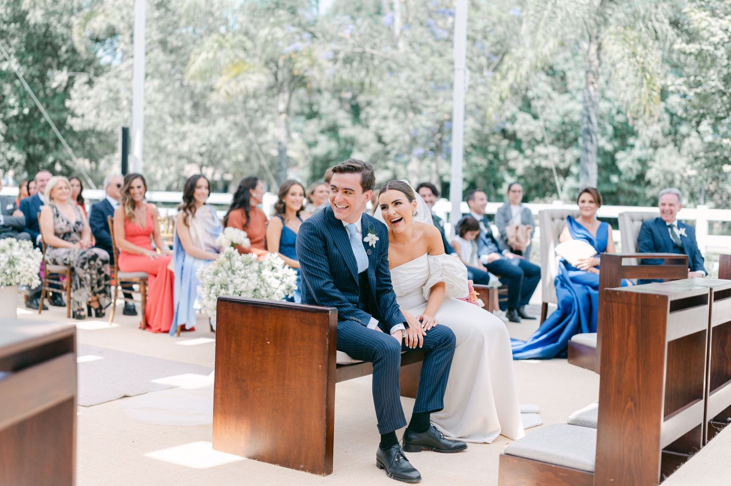 Bride and groom sit together on wooden pew during wedding ceremony with guests seated in background.