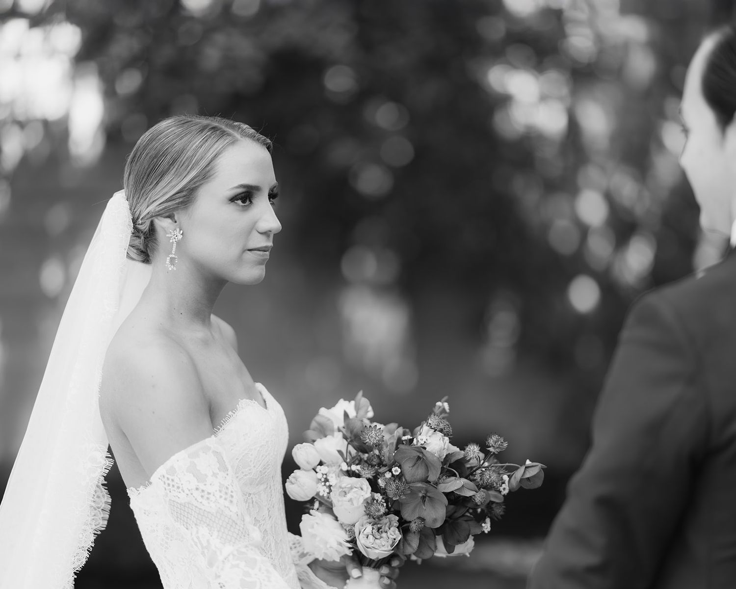Black and white portrait of a bride holding a bouquet gazing at her groom during the ceremony.