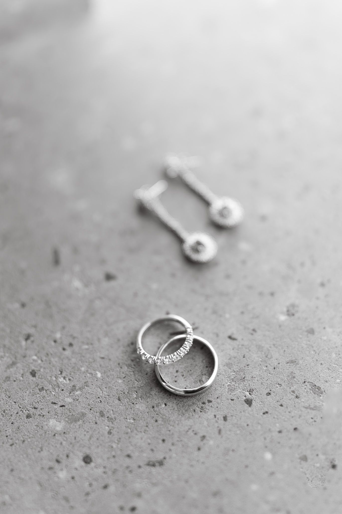 Black and white close-up of wedding rings and diamond drop earrings on a textured stone surface.