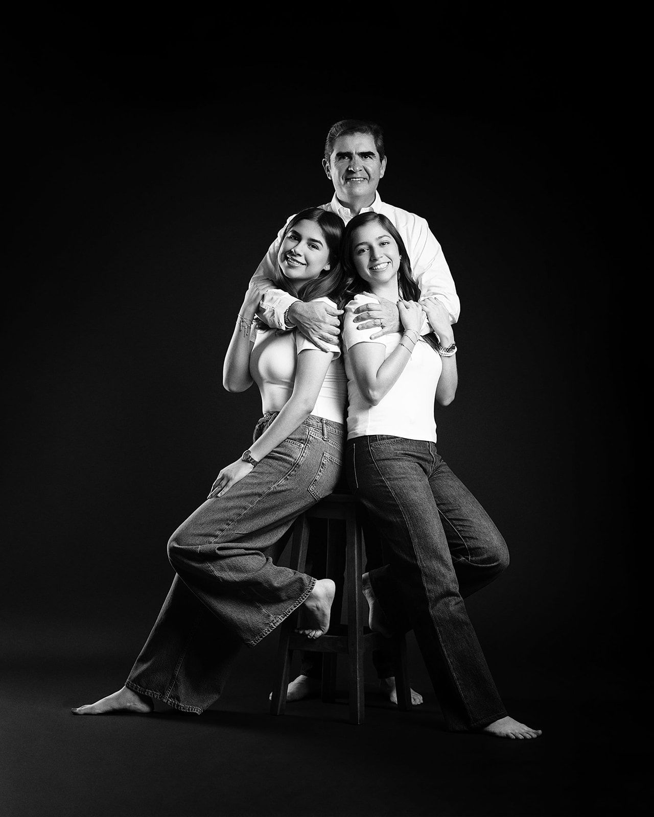 Black and white studio portrait of father seated with two daughters embracing him from both sides.