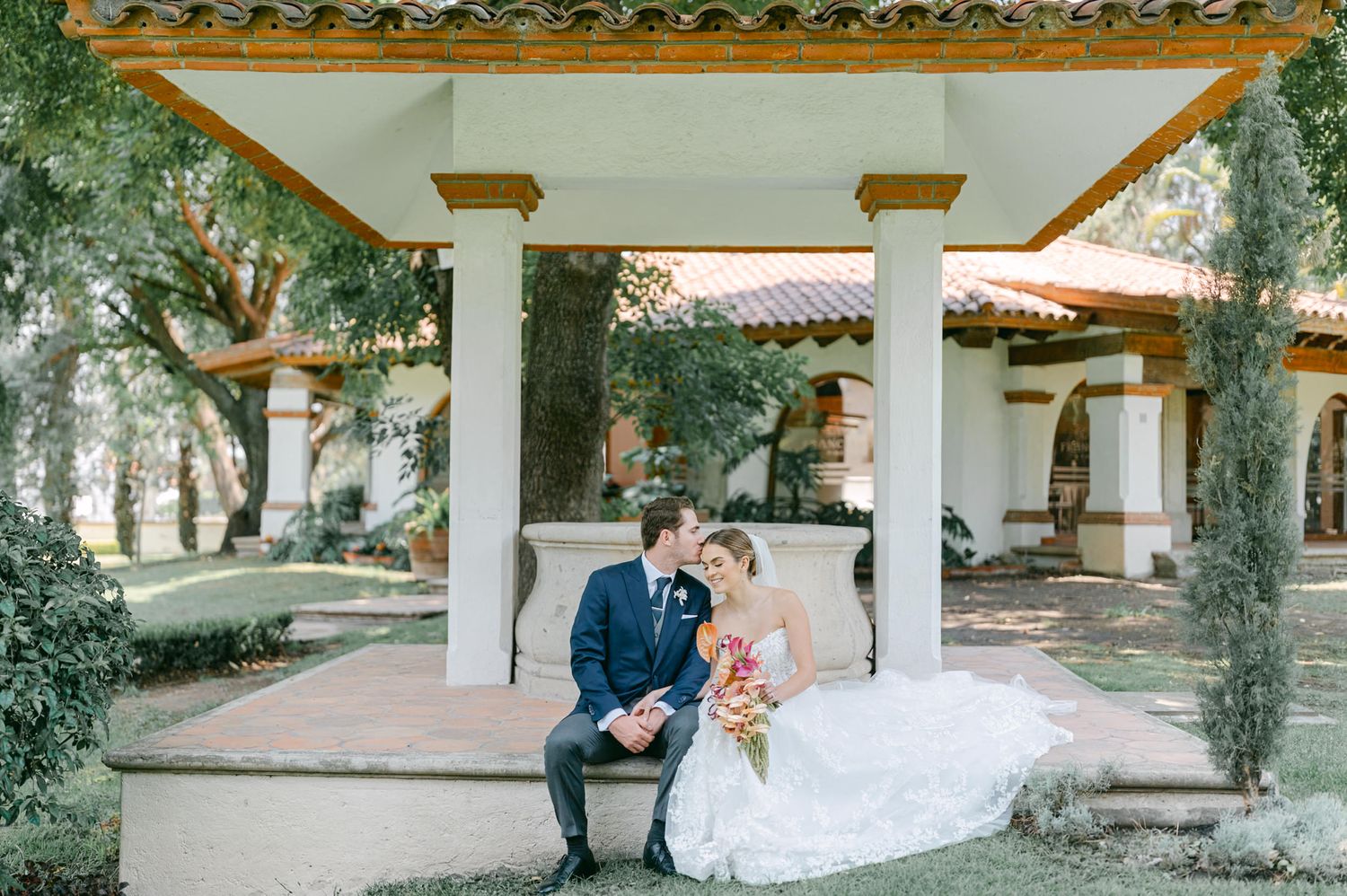 Bride and groom sharing a kiss while seated under elegant Spanish-style pavilion at wedding venue.