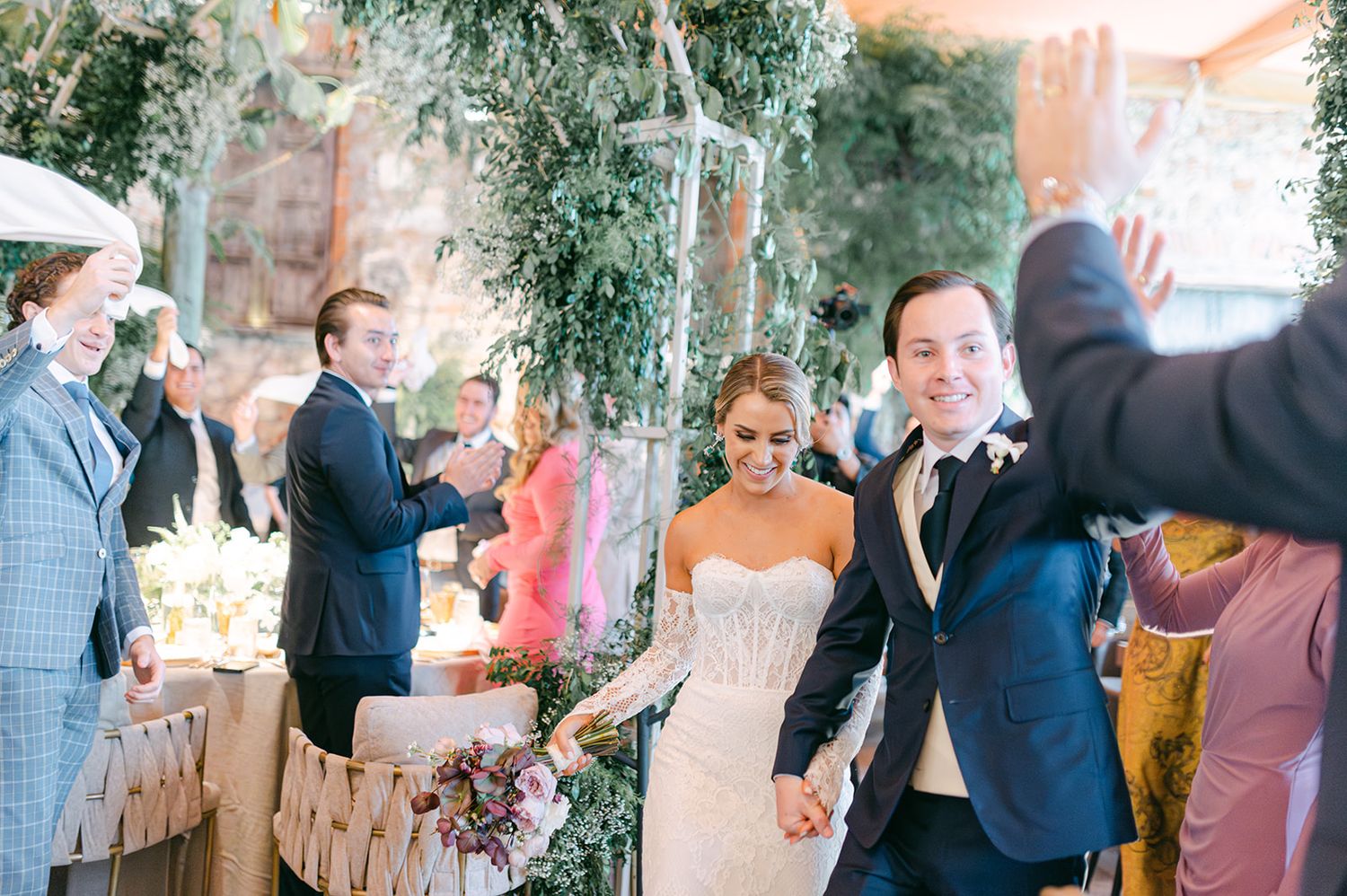 Smiling newlyweds walk hand in hand through applauding guests at lush garden wedding reception entrance.