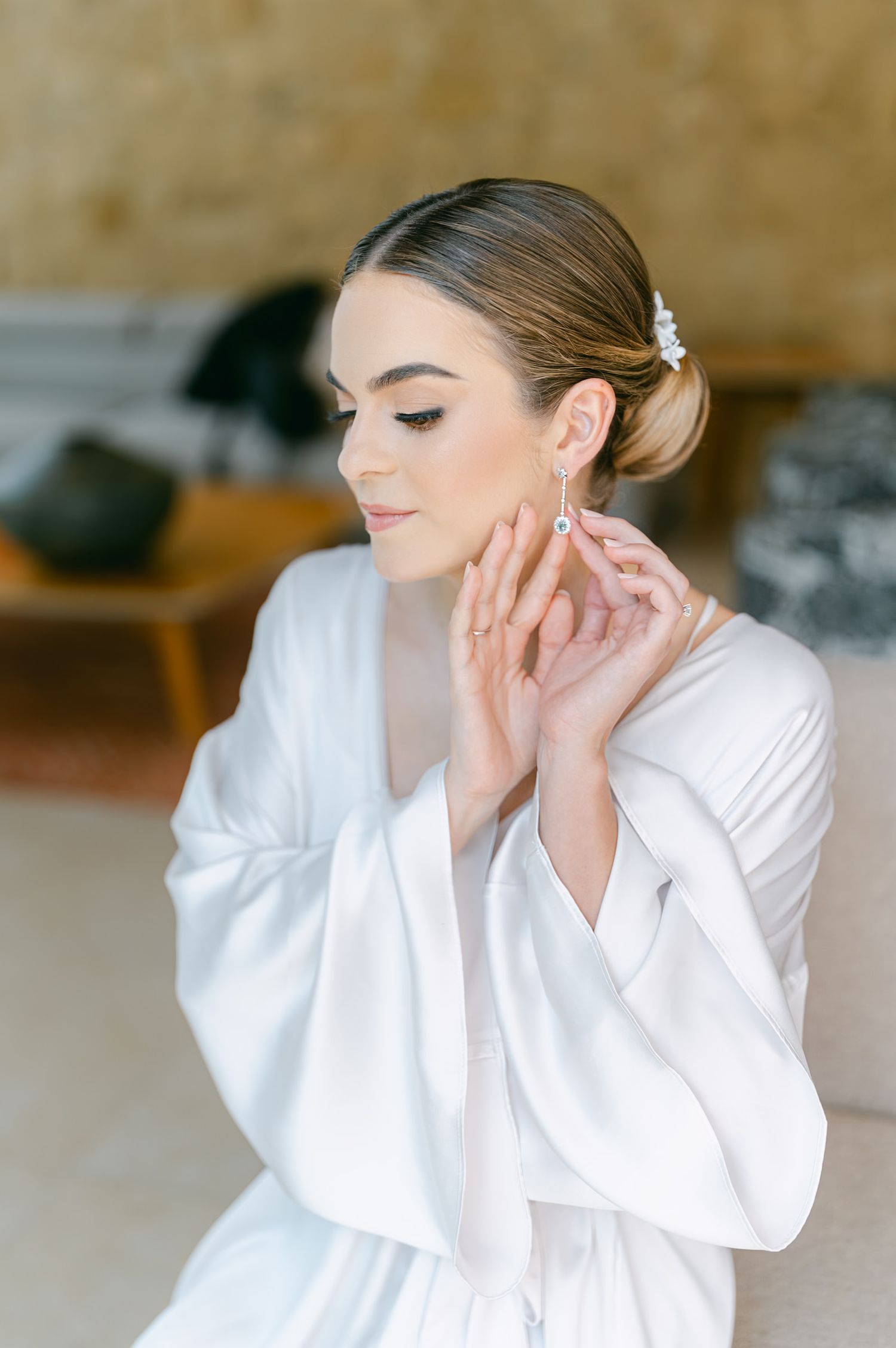 An elegant bride in a white silk robe delicately puts on a drop earring before her wedding.