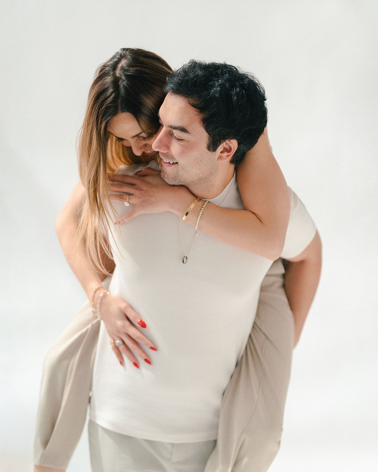 A smiling couple shares a joyful piggyback ride, woman in white top with red nails embracing man in beige outfit.
