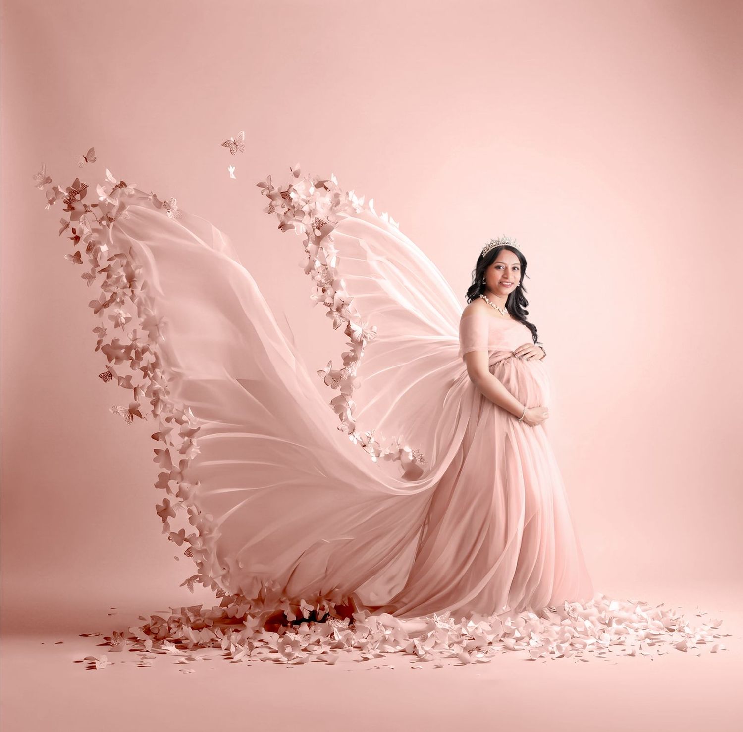 Pregnant woman in flowing pink gown with butterfly wings and floral details poses gracefully against a soft pink background.