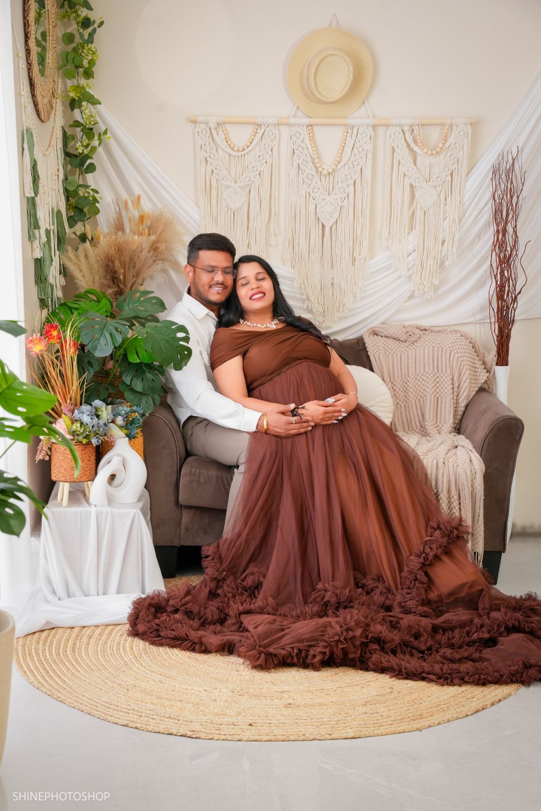 Couple poses for maternity photoshoot, woman in brown tulle gown, boho studio backdrop with macrame wall hanging.