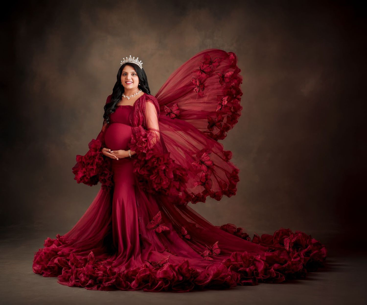 Pregnant woman in elegant red tulle butterfly gown with floral details and tiara poses for maternity portrait.