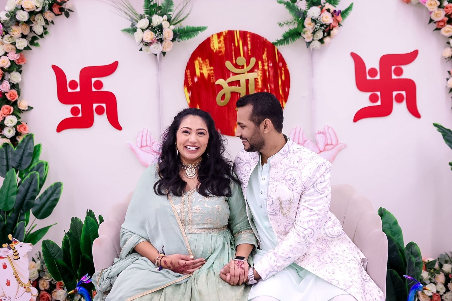 Couple shares intimate moment during maternity photoshoot with decorative floral and traditional Hindu backdrop.