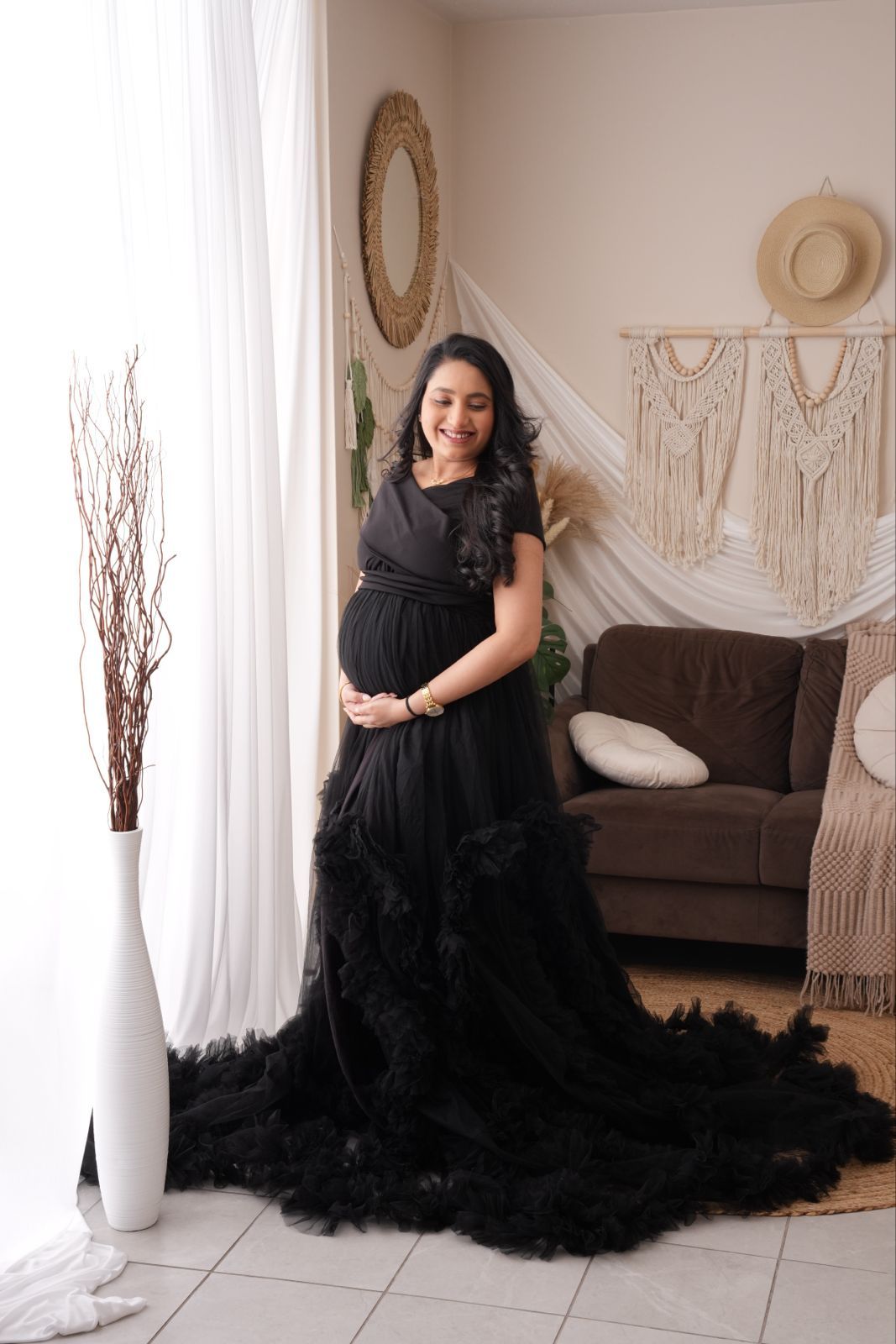 Pregnant woman in elegant black ruffled tulle maternity gown posing in boho-styled room with macrame wall decor.