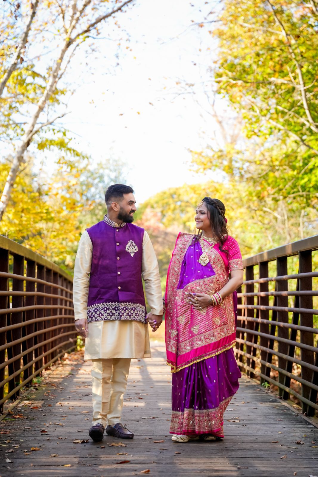 A couple in purple and pink Indian wedding attire walks hand in hand on a wooden bridge in autumn.