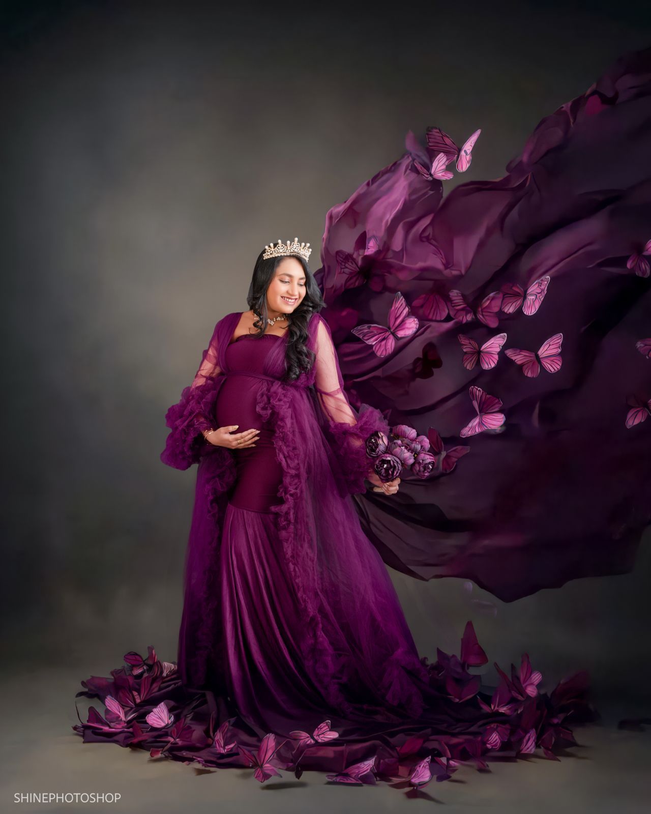 Pregnant woman in flowing purple gown with crown, surrounded by butterflies in elegant maternity photo shoot.