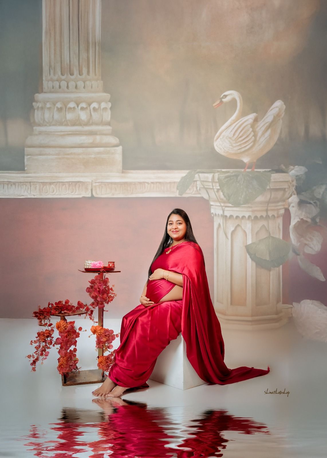 Artistic maternity portrait with red saree against classical architectural backdrop with swan decor.