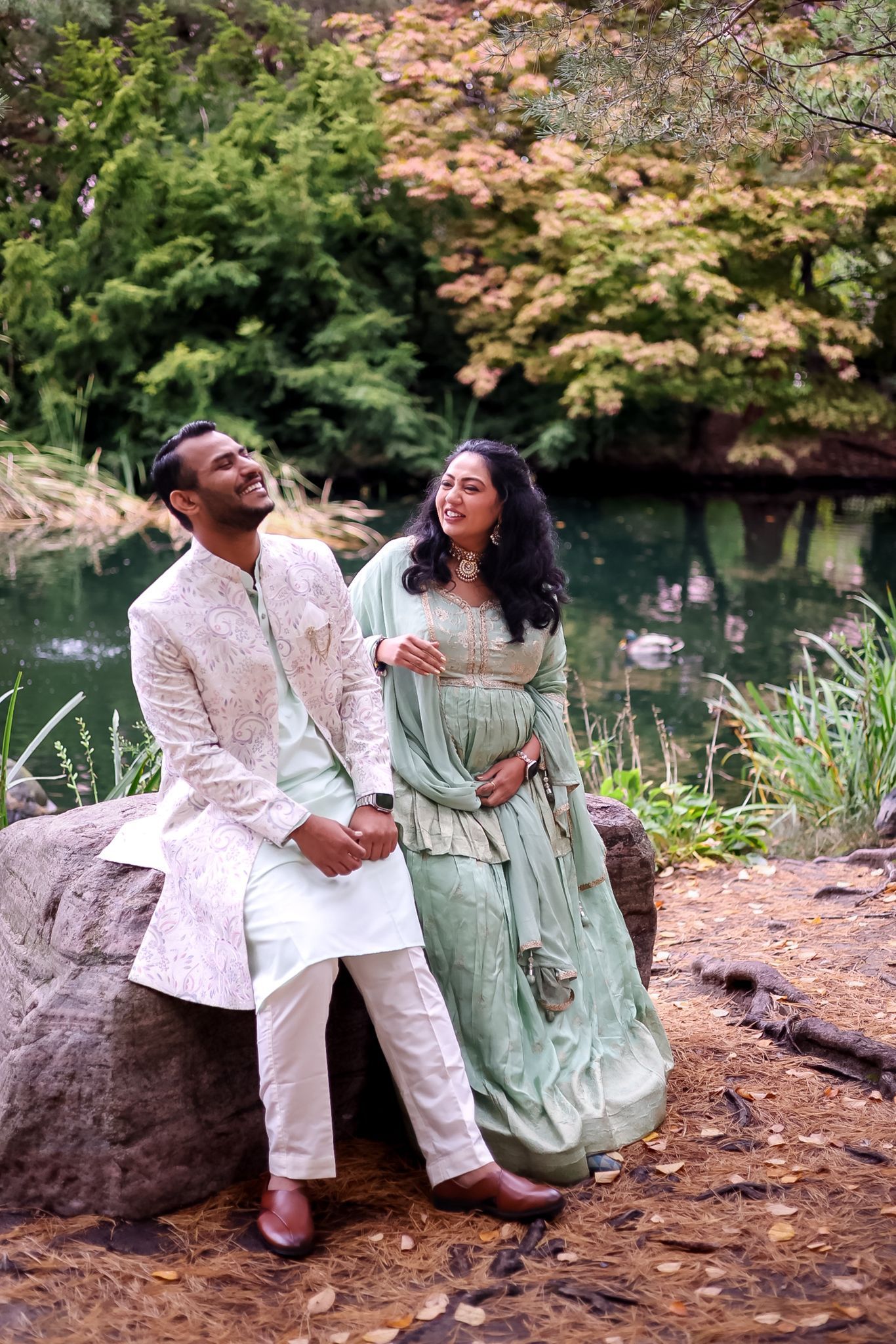 A couple shares a joyful moment while posing on rocks near a garden pond.