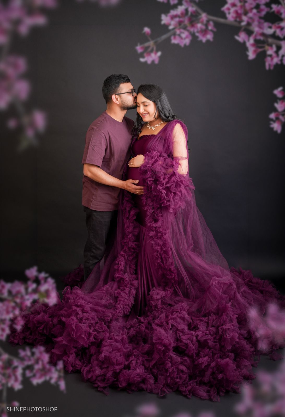 Couple in maternity photoshoot, woman in purple ruffle gown, man in mauve shirt, dark background with pink flowers.