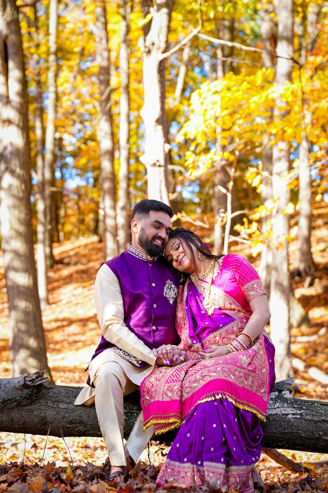 A couple in traditional Indian attire sits on a log surrounded by golden autumn foliage.