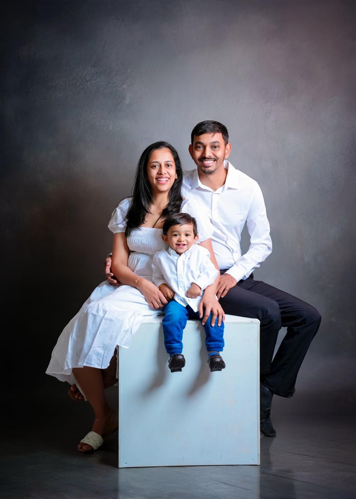 A family portrait showing a couple and young child sitting together on a white cube against a dark background.