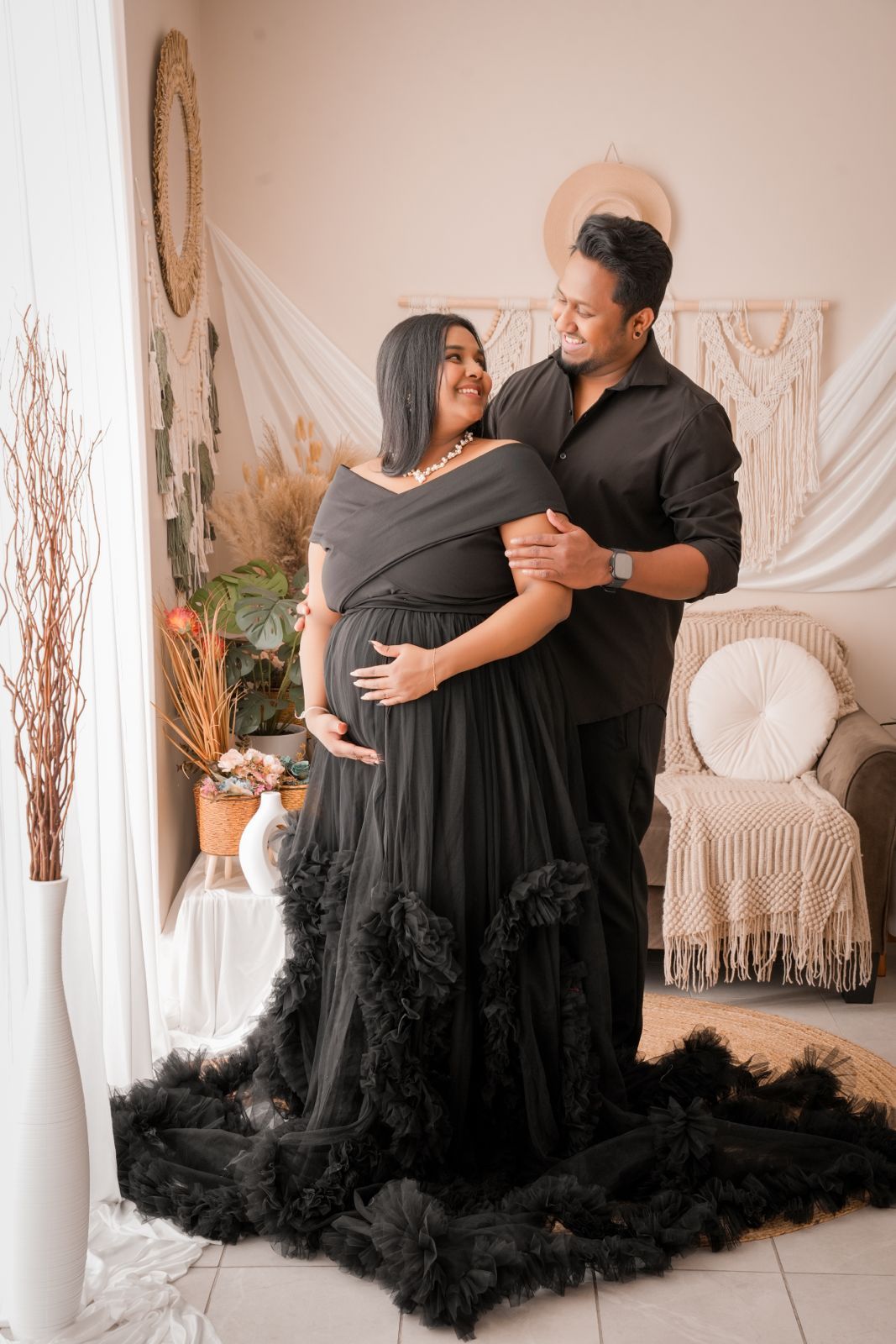 Couple in matching black outfits posing for maternity photo in boho studio, woman in ruffled gown cradling baby bump.