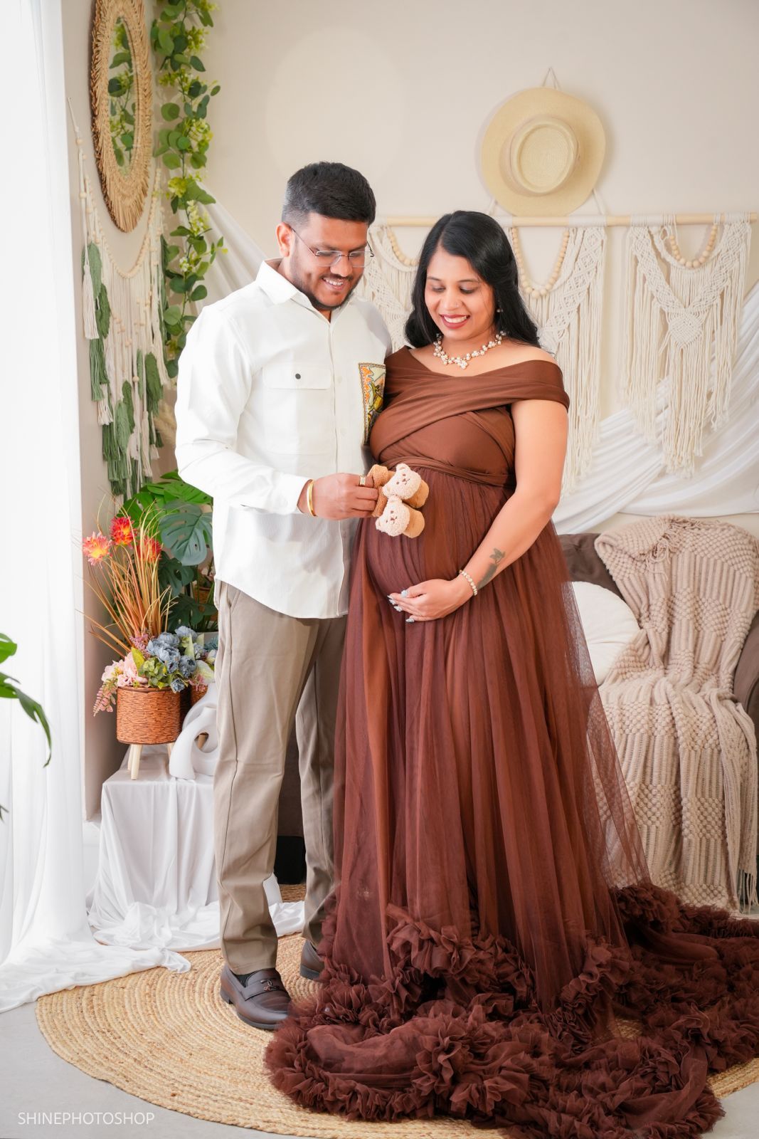 Couple in maternity photoshoot, woman in brown gown holding baby shoes, man in white shirt, boho studio backdrop.