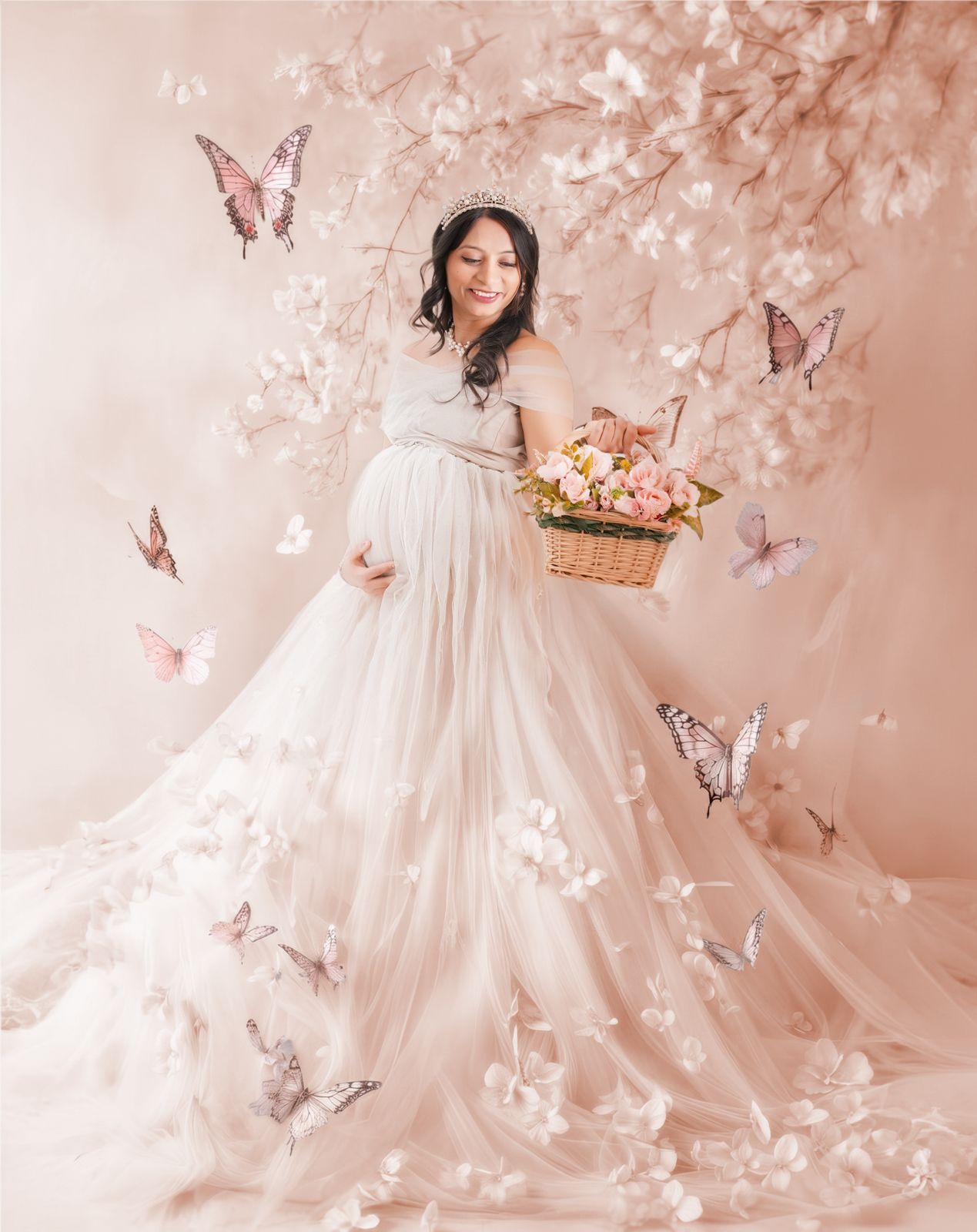 Pregnant woman in blush tulle gown holding flower basket, surrounded by butterflies and cherry blossoms in dreamy studio.
