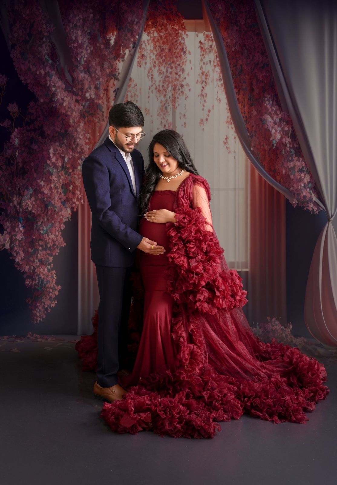 Couple in elegant maternity photoshoot, woman in red ruffled gown, man in navy suit, floral backdrop.