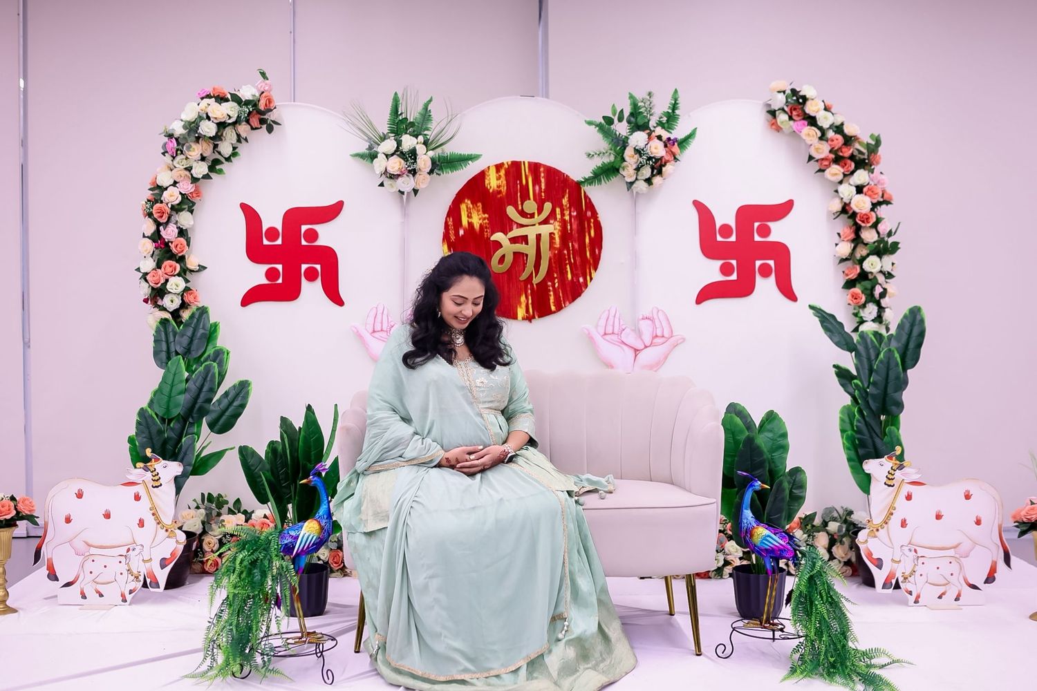 Couple poses in front of pink wall with floral decorations and red swastika symbols during baby shower celebration.