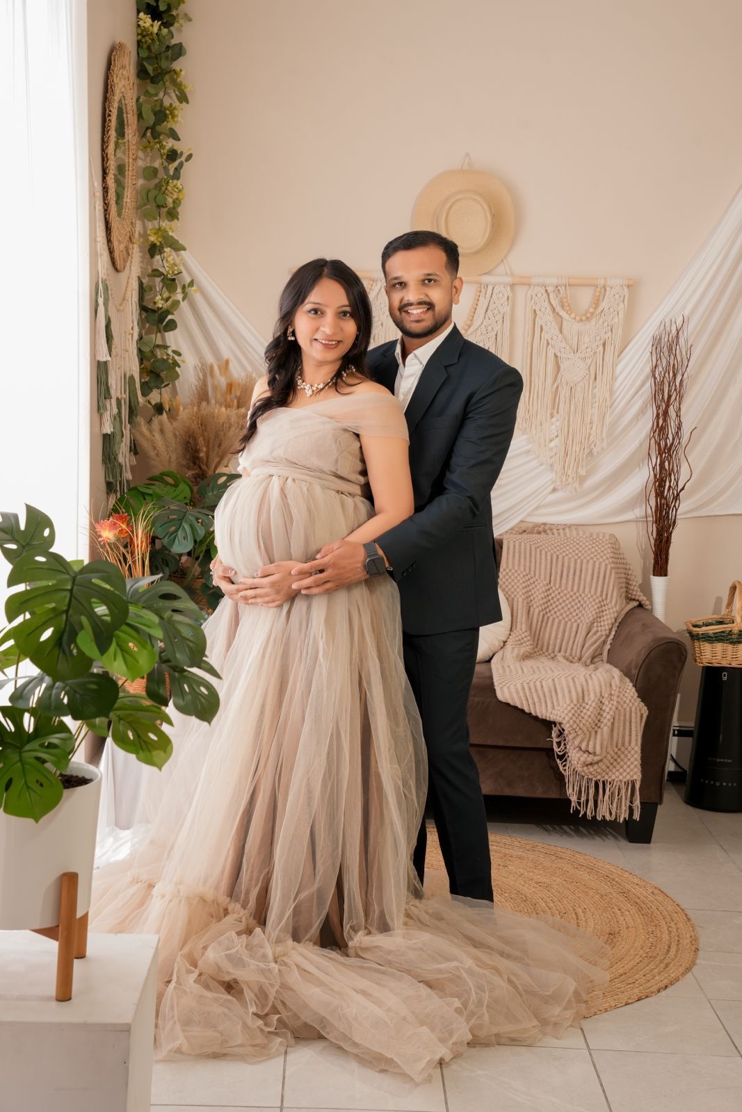Couple posing for maternity photo, woman in beige tulle gown cradling bump, man in black suit, boho studio backdrop.