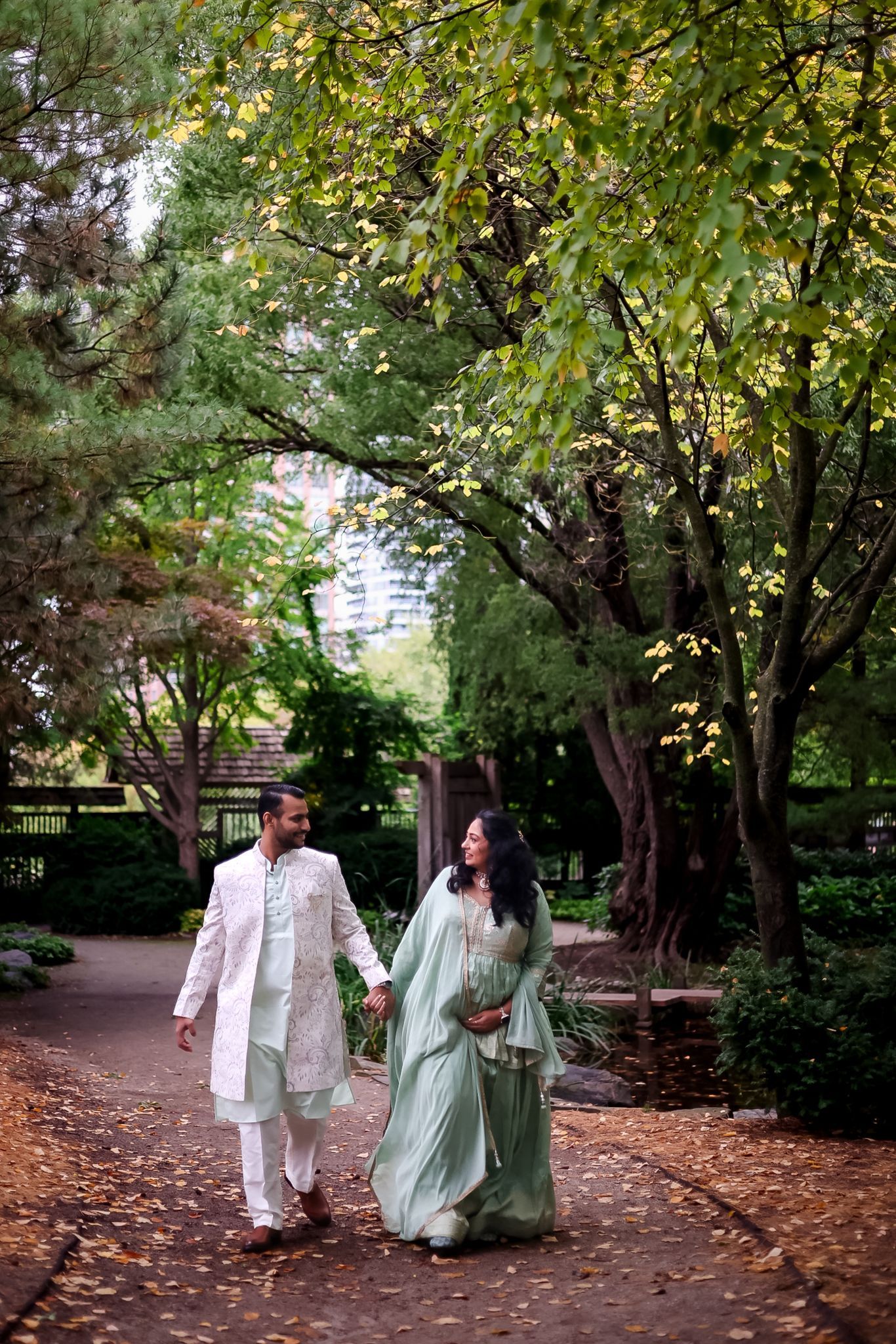 A couple strolls through a park pathway covered in fallen autumn leaves.