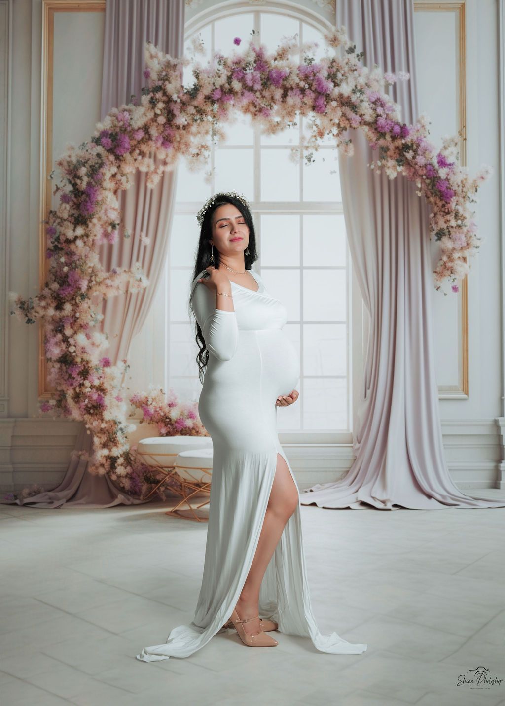 Elegant maternity photo with flowing white dress against floral arch and window backdrop.