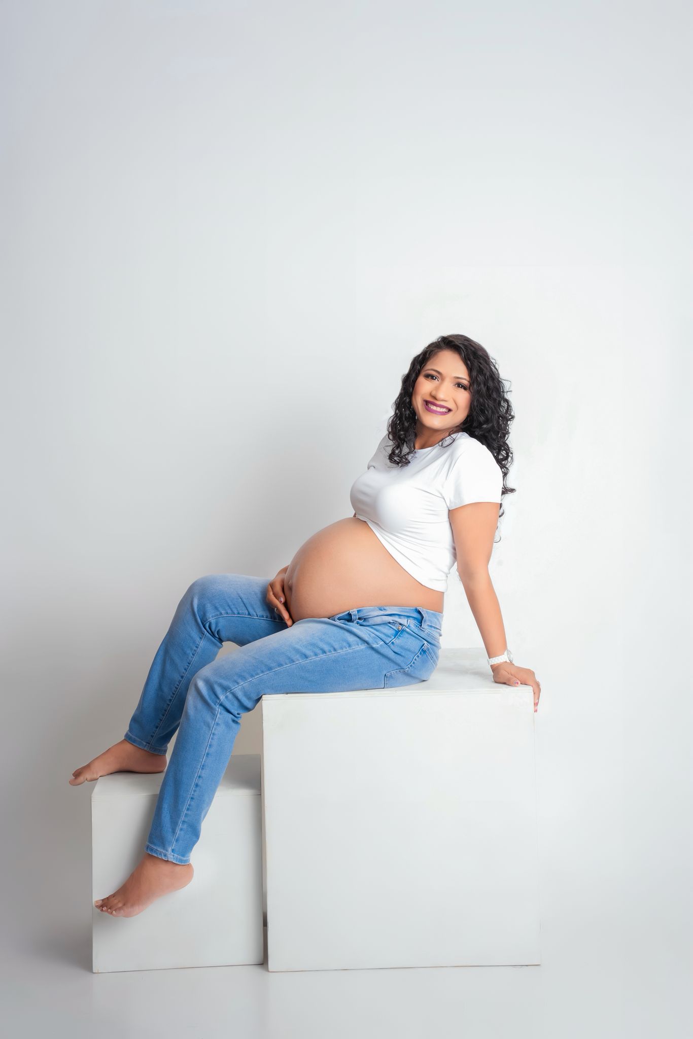 Pregnant woman in white top and jeans poses on white blocks during maternity photoshoot.