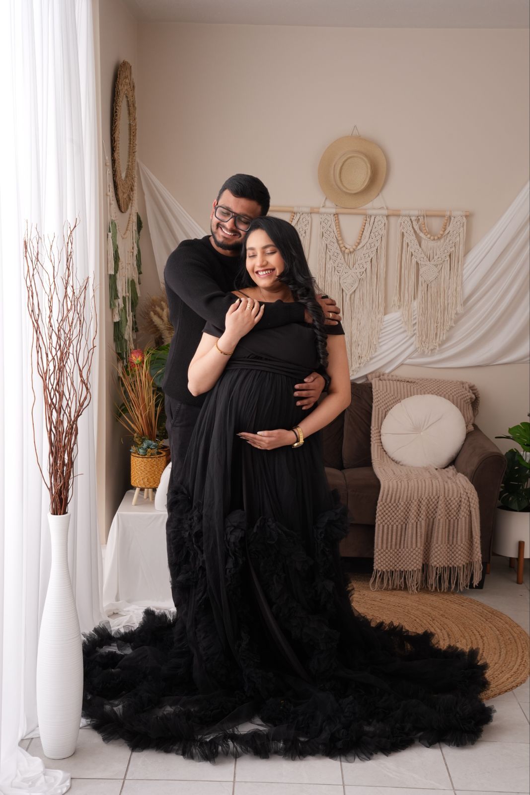 Couple in matching black outfits posing for maternity photo in boho studio, woman in ruffled gown cradling baby bump.