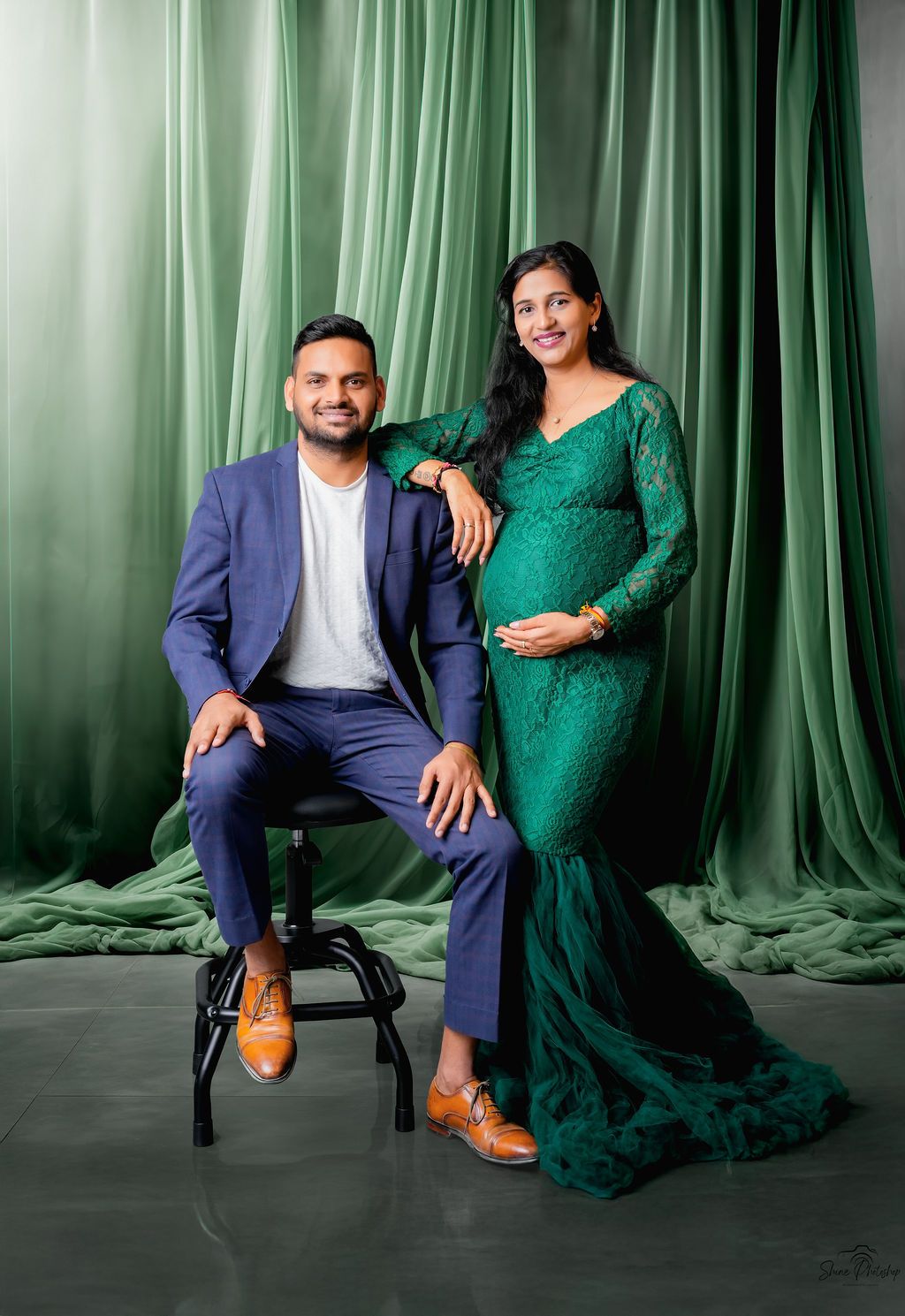 Elegant maternity photoshoot featuring a green lace gown and navy suit against emerald curtain backdrop.