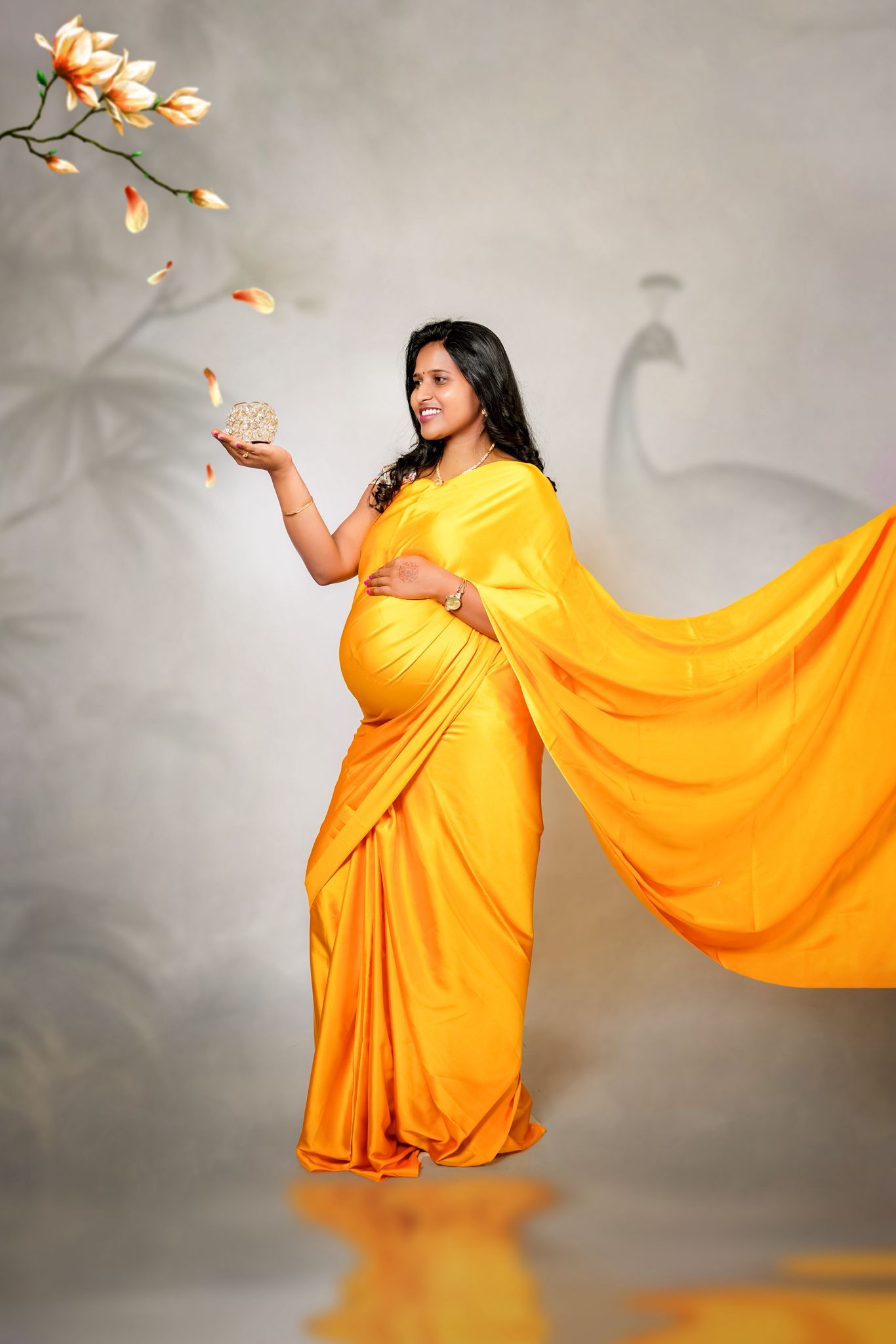 A woman in a flowing yellow sari poses elegantly against a gray backdrop with magnolia flowers in the frame.