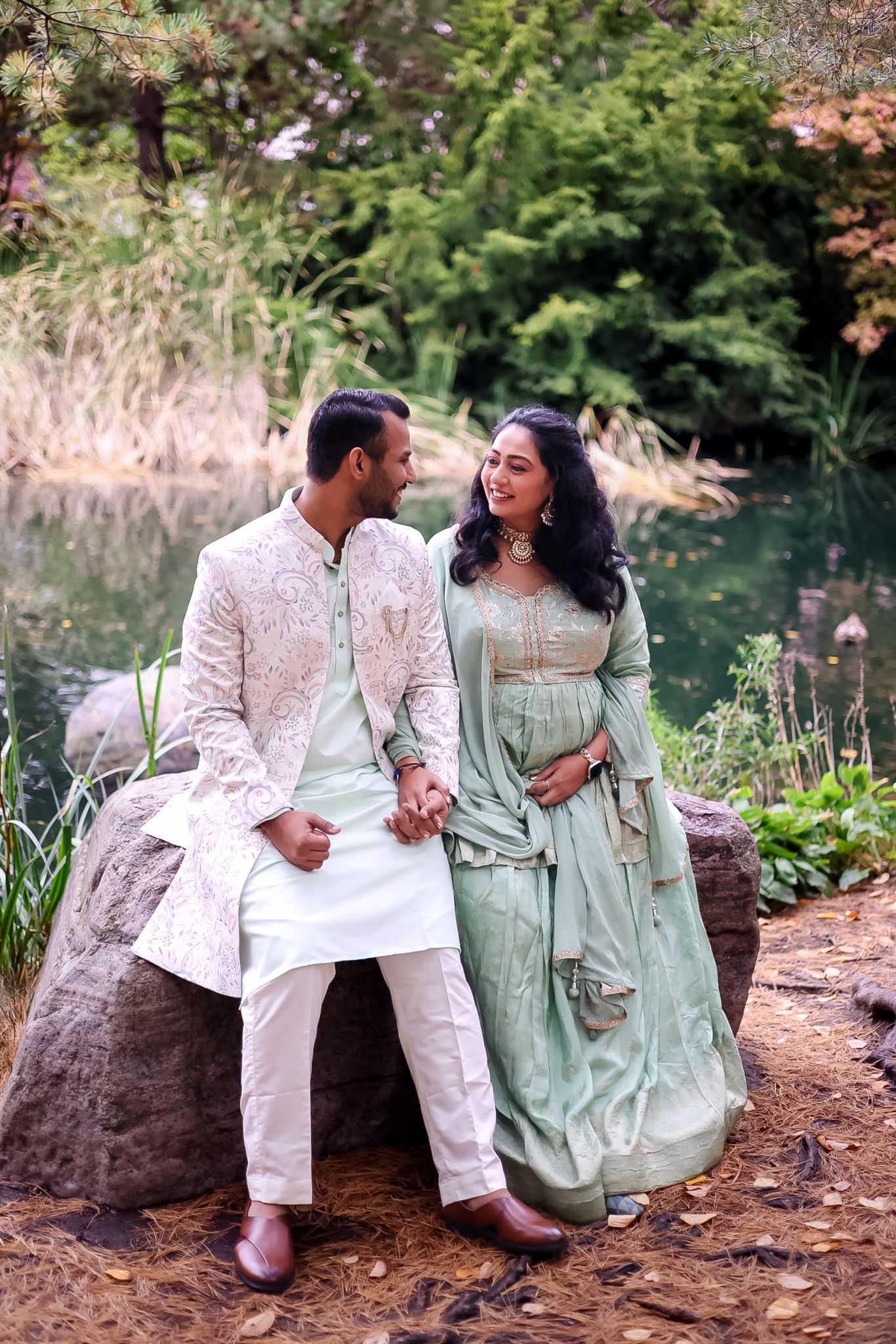 A couple in traditional Indian attire sits on rocks beside a serene pond during their photoshoot.
