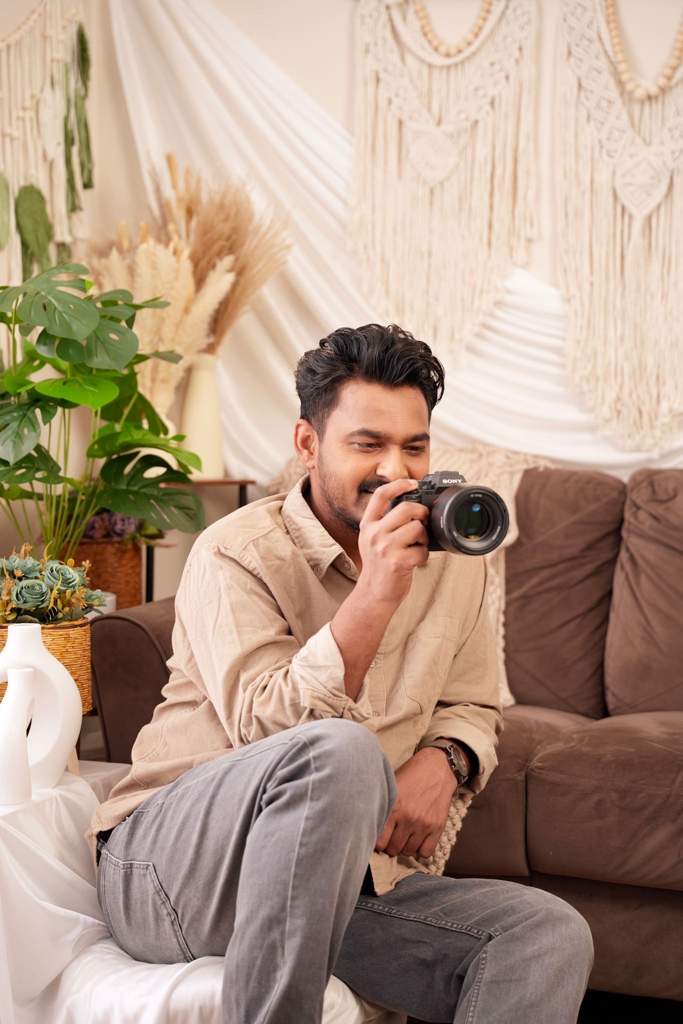 A photographer in casual attire sits on a brown couch examining their camera in a boho-styled room with macrame decor.