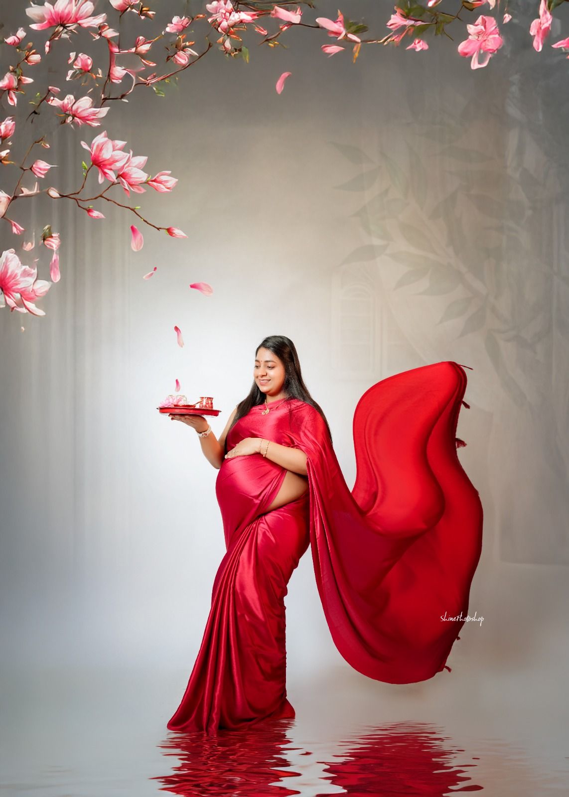 Dramatic maternity photo with flowing red saree and falling cherry blossoms.