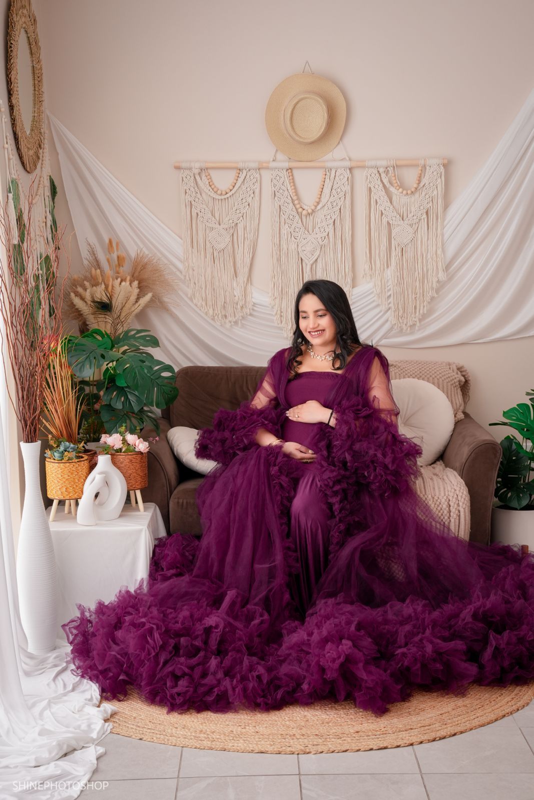 Pregnant woman in a flowing purple tulle gown poses gracefully in a boho-styled photography studio.