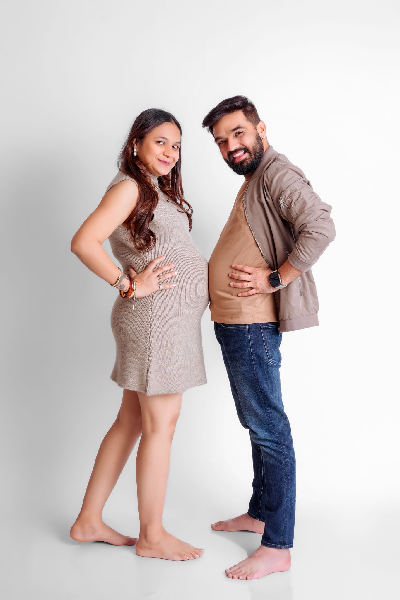 Playful barefoot maternity photo with couple in casual attire against white background.