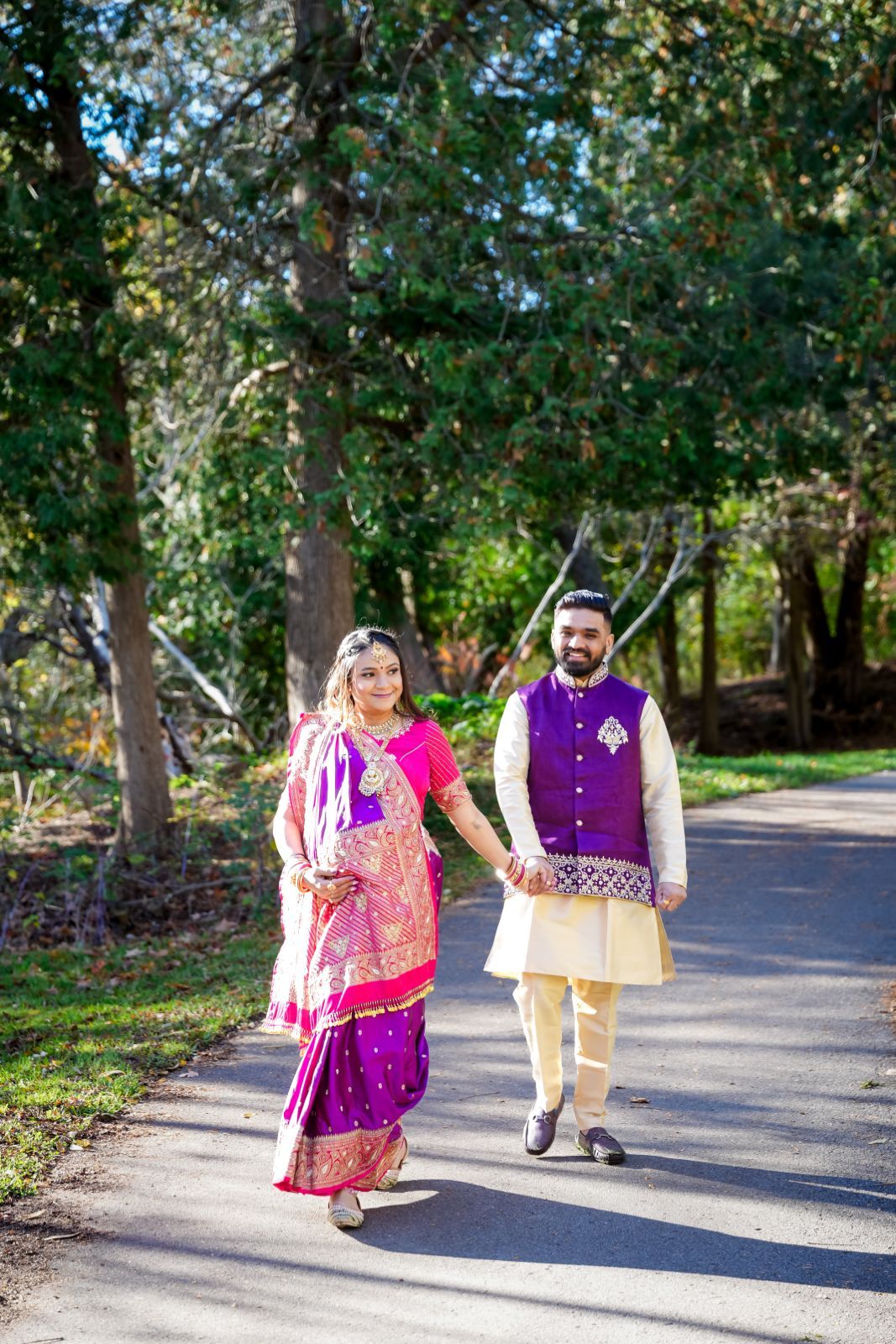 A couple in vibrant Indian formal wear strolls along a tree-lined path.