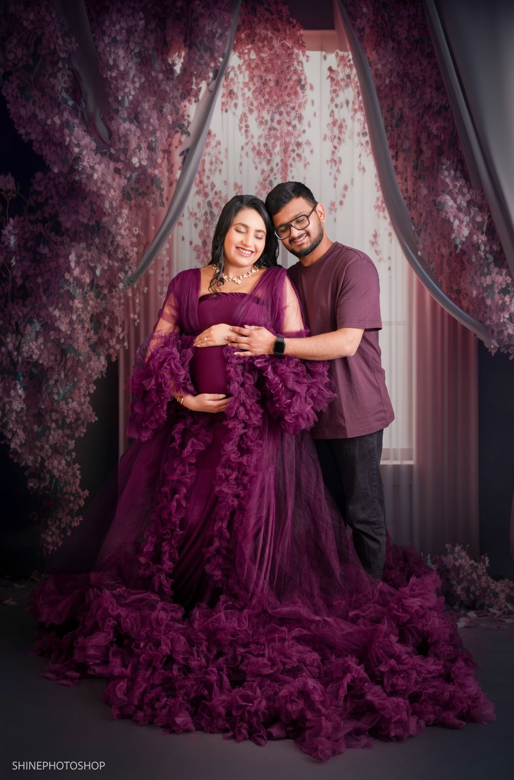 Couple in maternity photoshoot, woman in purple ruffled gown, man in maroon shirt, floral backdrop.