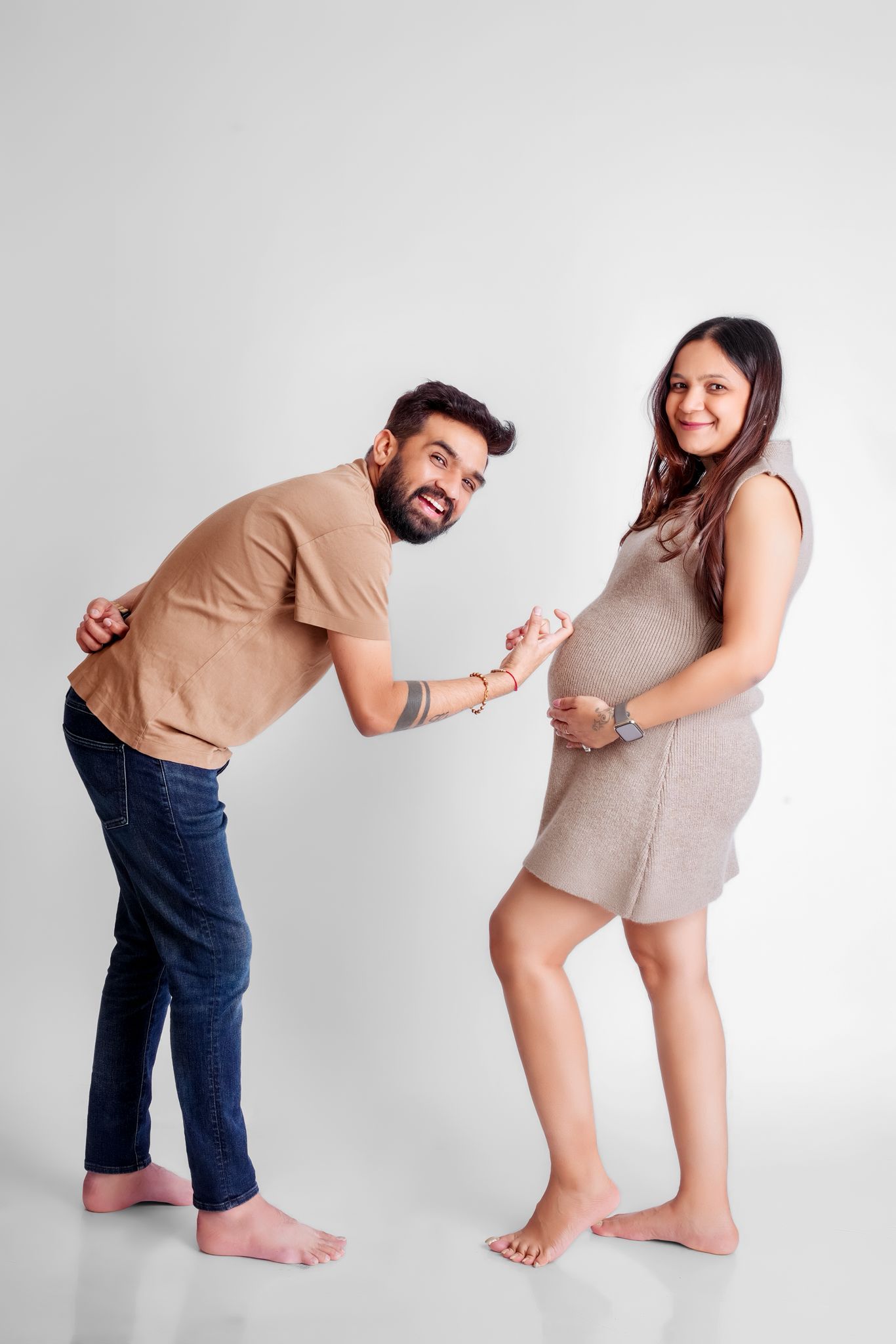 Fun maternity pose with barefoot couple in casual wear against white backdrop.