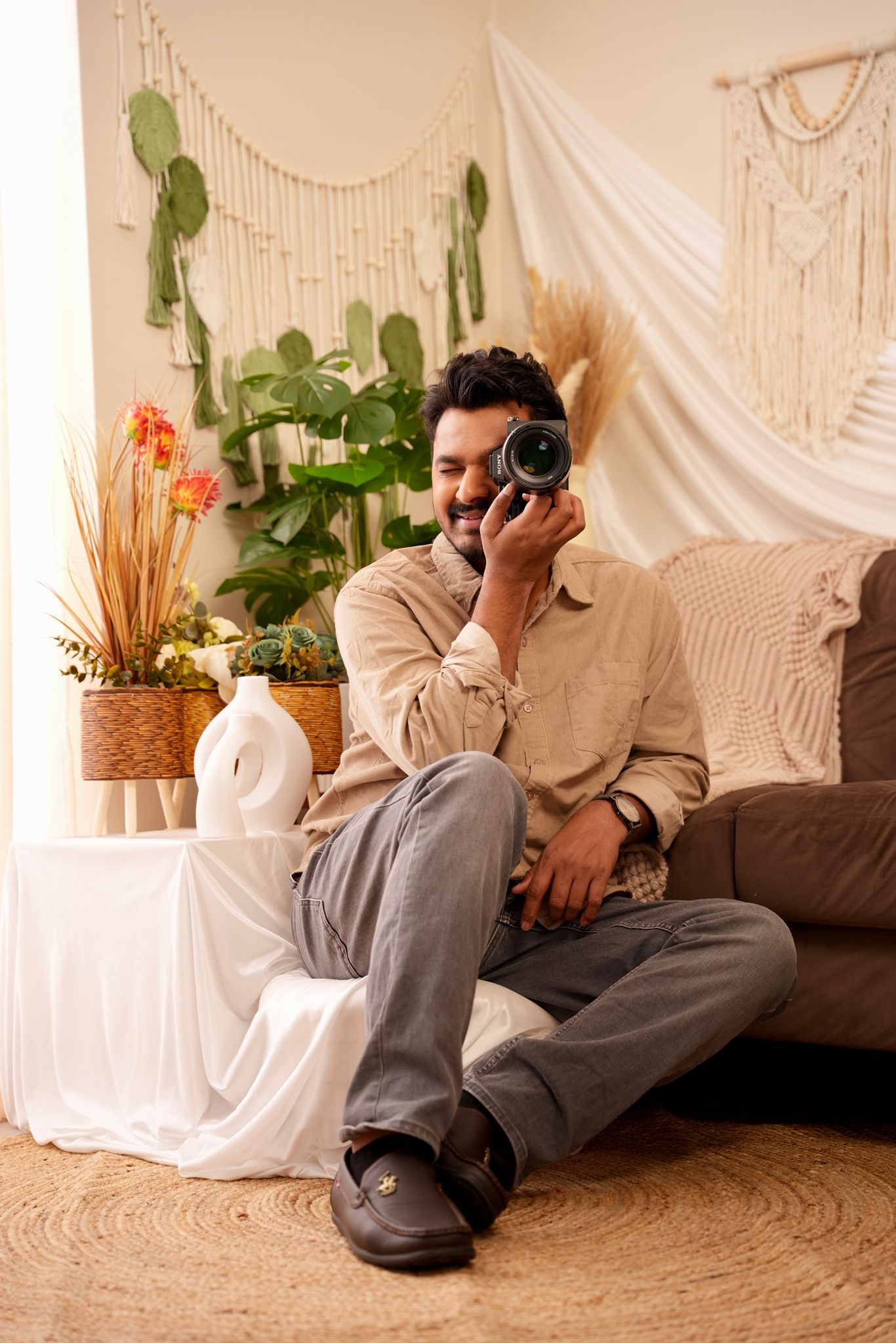 Person in beige sweater and gray pants sits on couch with camera in bohemian styled room with macrame and plants.