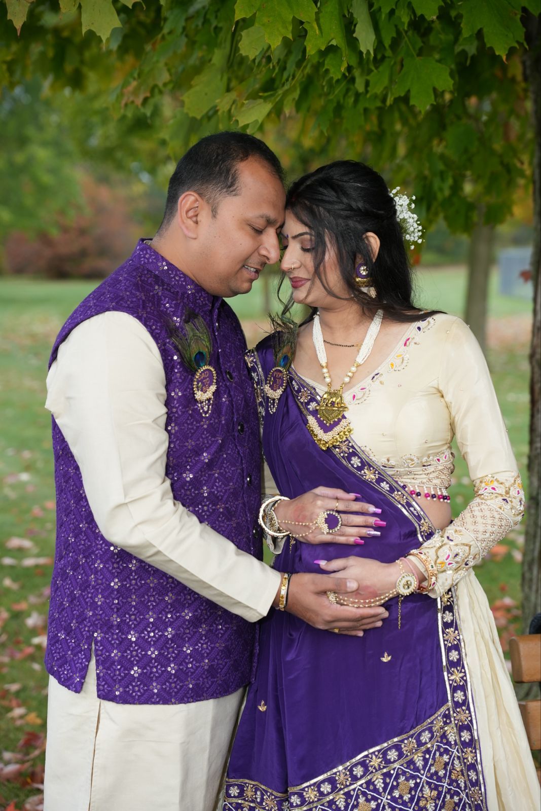 A couple in matching purple and cream traditional outfits shares an intimate moment under green leaves.