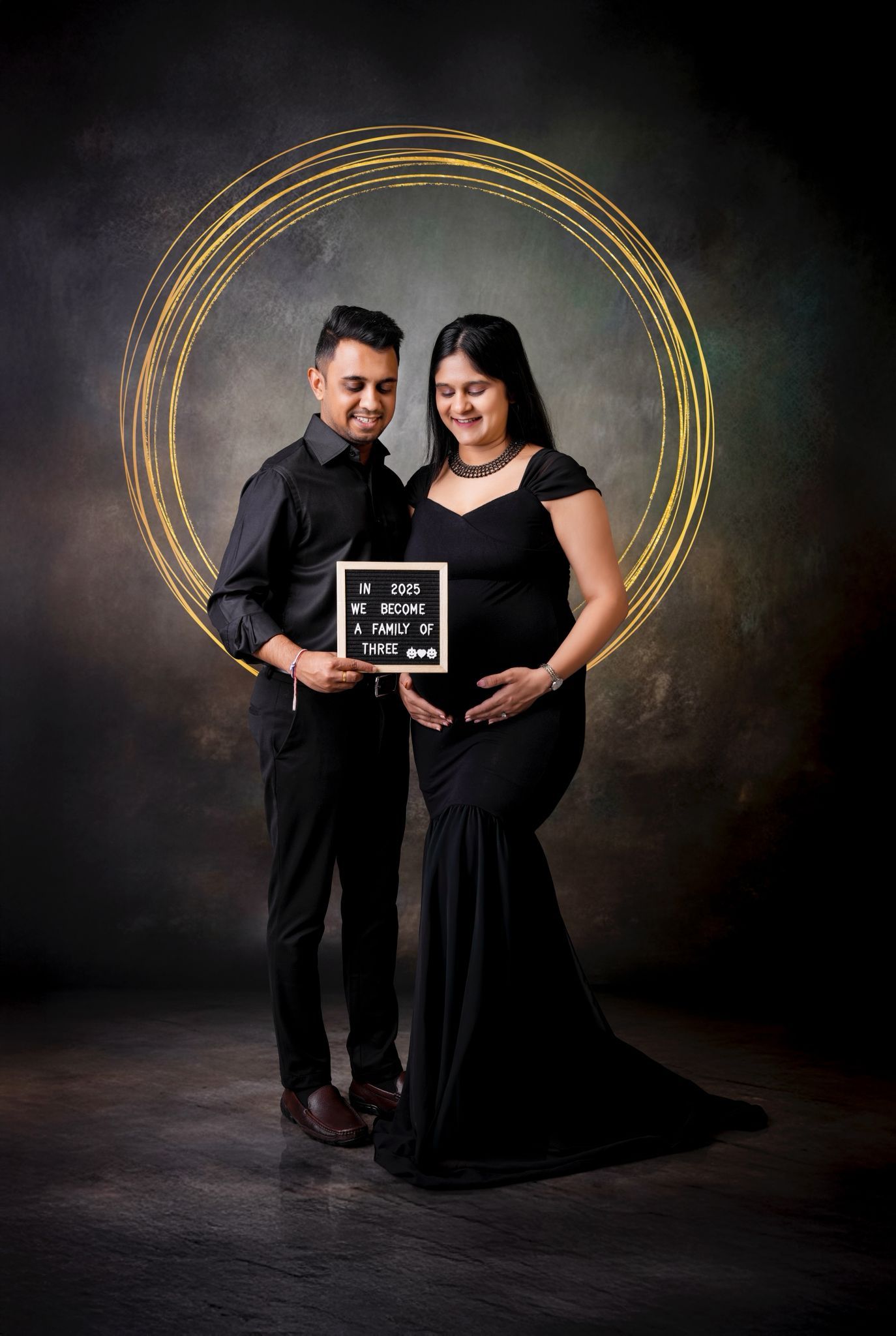 Dramatic maternity portrait with couple in black attire against dark background with gold circles.
