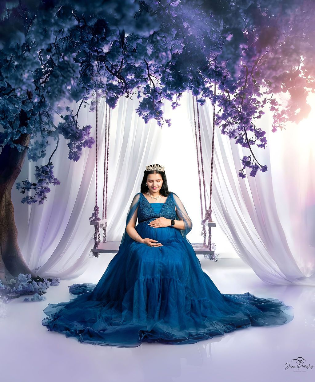 Maternity portrait on swing with blue gown against purple floral and white curtain backdrop.