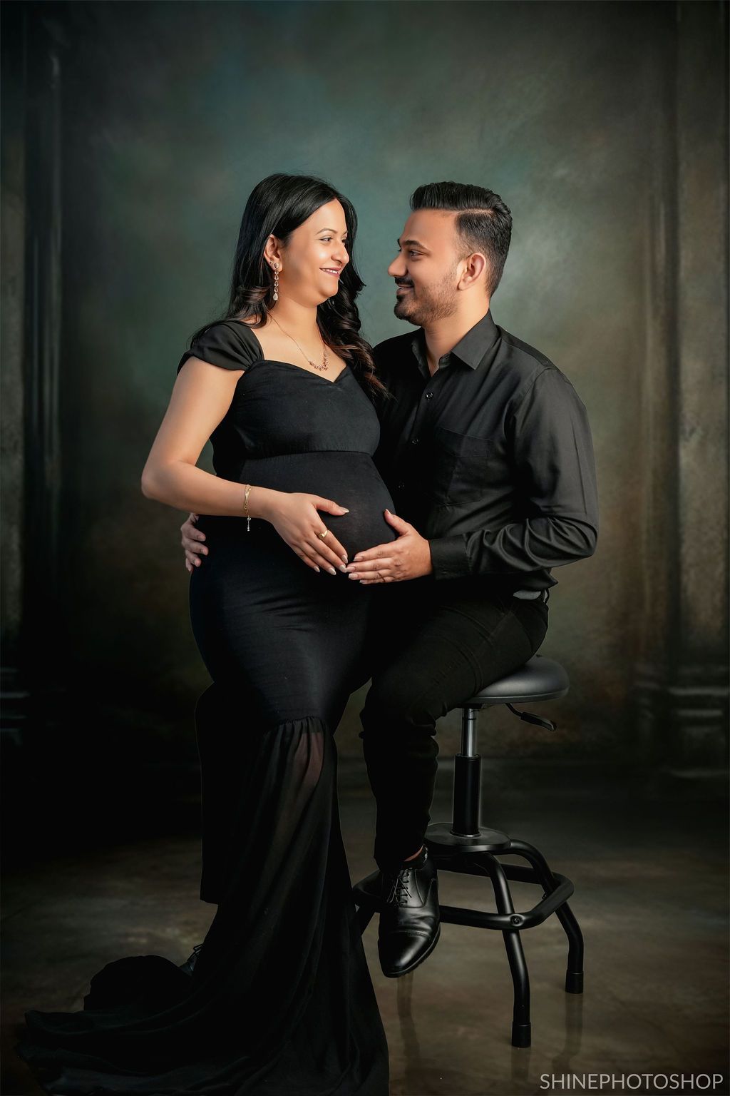 Intimate maternity session with couple in black attire against dark moody background.